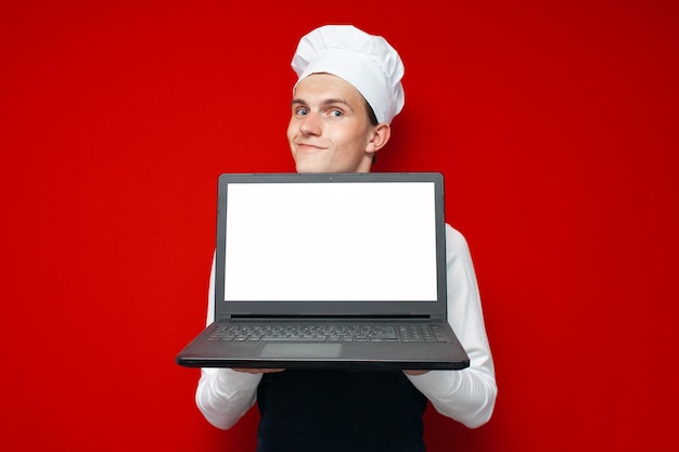 Cook shows blank laptop screen on red isolated background guy in chef uniform