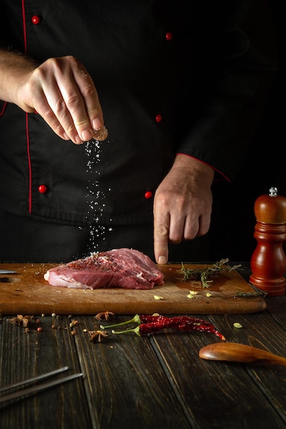 The cook sprinkles salt into meat steak for cooking for barbecue lunch Working environment on the kitchen table with fragrant rosemary and garlic