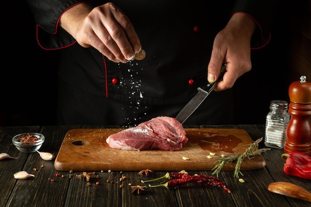 The cook sprinkles salt on the raw meat Preparing beef steak before baking Working environment in a restaurant or cafe kitchen