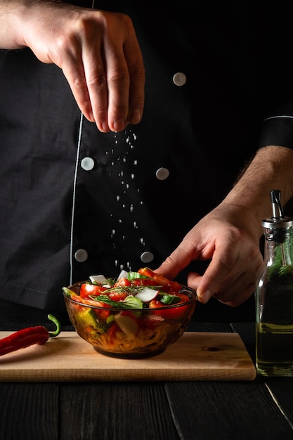 The cook sprinkles salted fresh vegetable salad in a plate on a wooden table