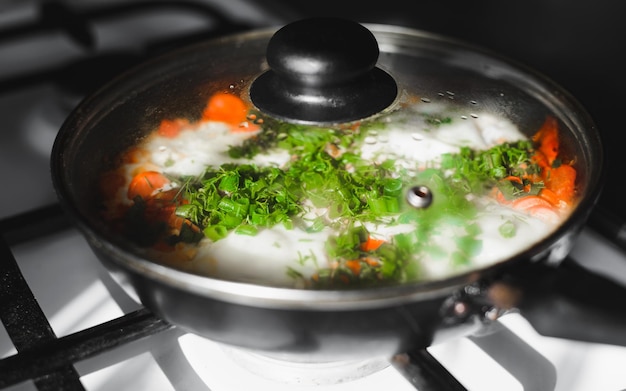 Cooked food in closed frying pan on gas stove closeup Fried eggs with tomatoes and onions shakshuka Selective focus on green onion