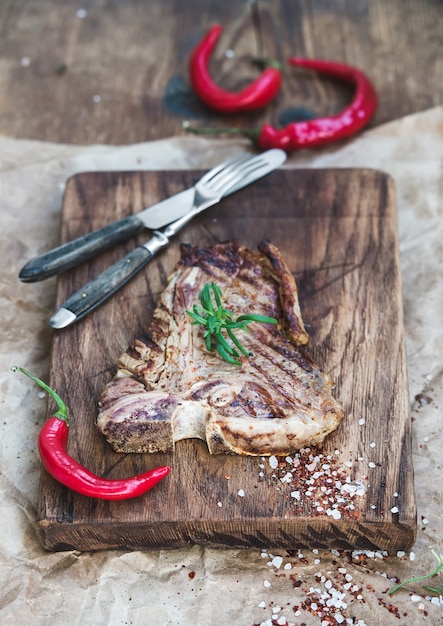 Cooked meat t-bone steak on serving board with red chili peppers, spices, fresh rosemary over oily craft paper and rustic wooden surface