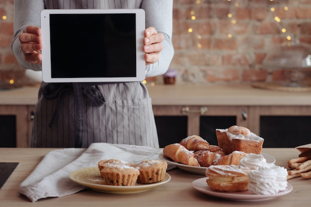 Photo cooking blog. homemade sweet bakery. man in apron with black screen tablet. fresh cakes and pastries assortment.