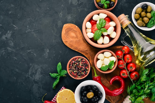 Cooking a caprese salad Mozzarella cheese cherry tomatoes olives basil leaves oil pepper On a black stone table Free space for text