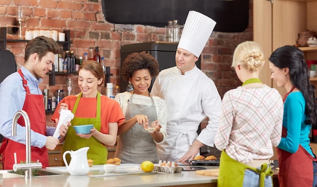 cooking class, culinary, bakery, food and people concept - happy group of friends and male chef cook baking in kitchen