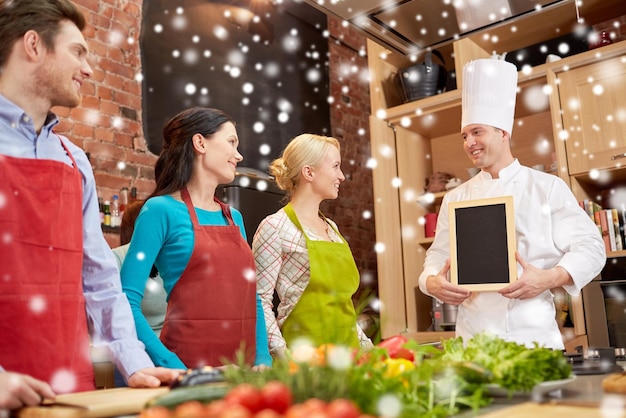 cooking class, culinary, food and people concept - happy friends and chef cook with blank menu chalk board in kitchen over snow effect