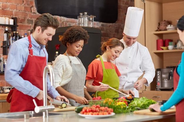 cooking class, culinary, food and people concept - happy group of friends and male chef cook cooking in kitchen