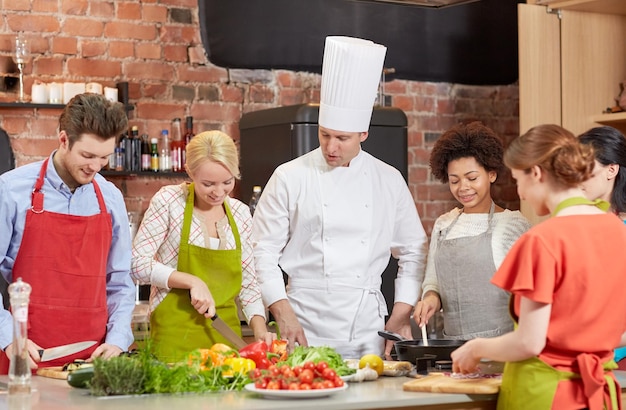 cooking class, culinary, food and people concept - happy group of friends and male chef cook cooking in kitchen