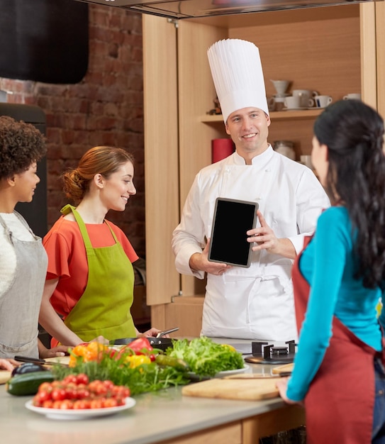 cooking class, culinary, food, technology and people concept - happy women with chef cook showing blank tablet pc screen in kitchen