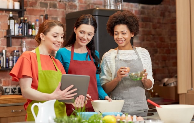 cooking class, friendship, food, technology and people concept - happy women with tablet pc computer in kitchen