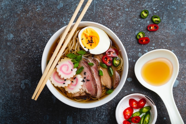 Cooking Japanese noodle soup ramen. Ramen with sliced pork, narutomaki, egg and kitchen spoon with broth on rustic stone background. Making traditional dish of Japan, top view, close-up, concept