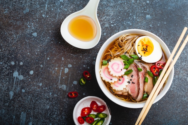 Cooking Japanese noodle soup ramen. Ramen with sliced pork, narutomaki, egg, kitchen spoon with broth on rustic stone background. Making traditional dish of Japan, top view, close-up. Space for text