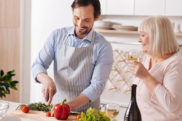 Cooking for my mother. Cheerful smiling mature son standing in the kitchen and cooking vegetarian salad while his aged mother drinking wine