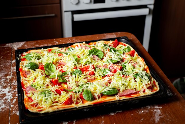 Cooking pizza with sausage tomatoes green leaves closeup on a baking sheet in the home kitchen