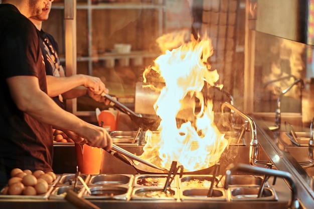 Cooking process in an Asian restaurant. Cook is fry vegetables with spices and sauce in a wok on a flame.