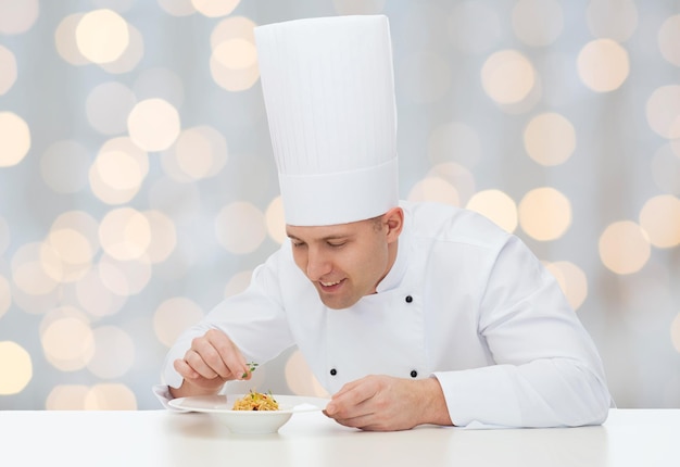 cooking, profession, haute cuisine, food and people concept - happy male chef cook decorating dish over holidays lights background