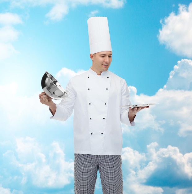 cooking, profession and people concept - happy male chef cook opening cloche cover over blue sky with clouds background