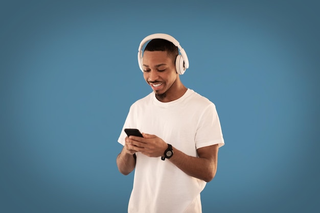 Cool african guy using wireless headphones and smarphone on blue