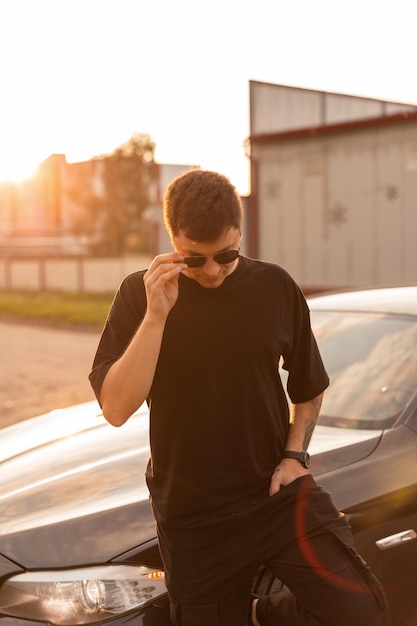 Cool fashion hipster man with sunglasses in black tshirt and jeans stands near a car at the sunset