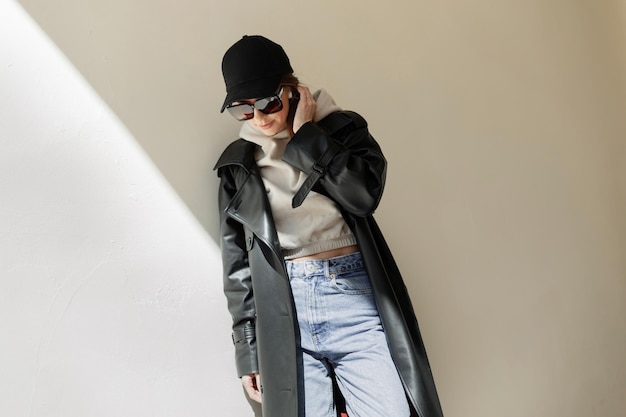 Cool fashion street beautiful woman hipster with trendy sunglasses and black cap in casual urban outfit with coat hoodie and jeans stands near a wall