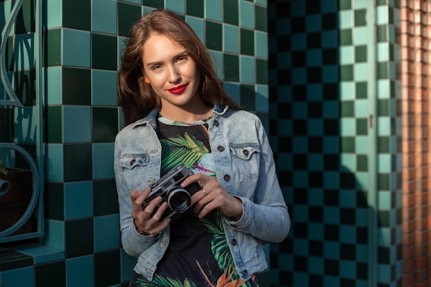 Cool girl model with retro film camera wearing a denim jacket, dark hair outdoors over city wall in a cage background. Having fun in the city with camera, travel photo of photographer.