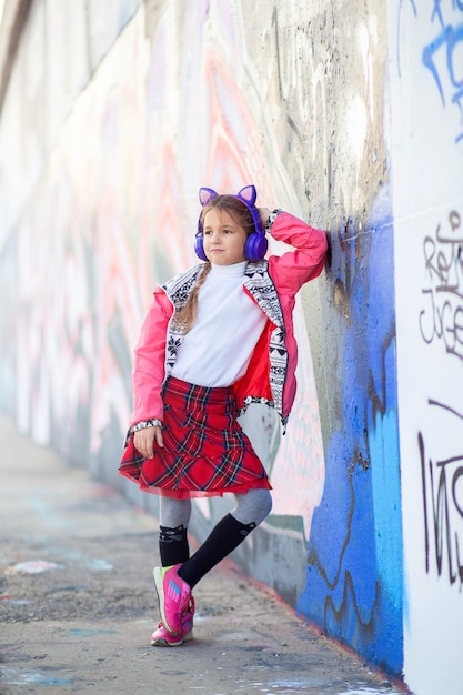 Cool girl in wireless headphones with ears poses near the city wall with graffiti.