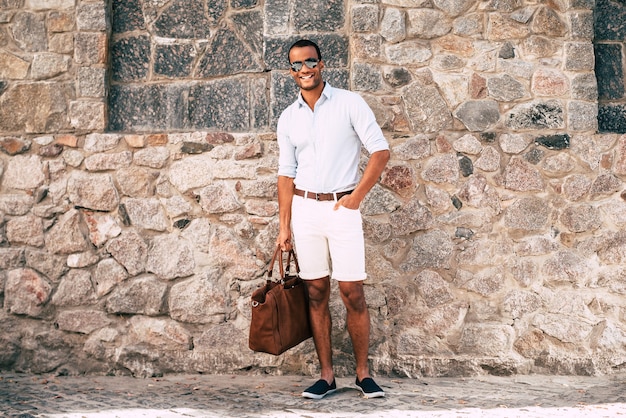 Cool and handsome. Full length of cheerful young African man in smart casual clothes carrying bag and looking at camera while standing against the stoned wall outdoors