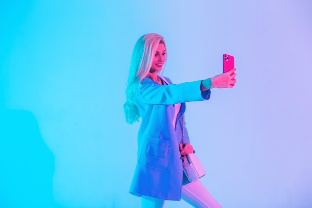 Cool happy young business woman in bright fashionable clothes with blazer takes selfie photo on phone in studio against neon pink light background