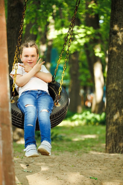 Cool little girl swinging on car rubber wheel with crossed arms in playground in green park Summer holidays in camp tourist center Playing outdoors sport activity and healthy lifestyle