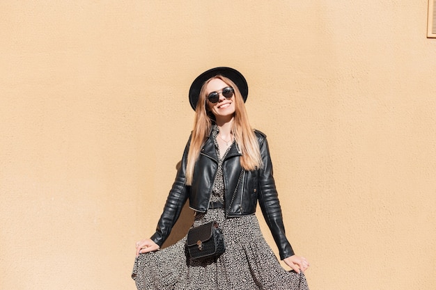 Cool pretty woman with smile in fashion look with leather jacket, vintage dress, sunglasses, hat and stylish handbag poses near a yellow wall outdoors