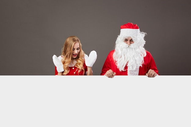 Cool Santa Claus and young beautiful mrs. Claus in sunglasses stand behind a white canvas