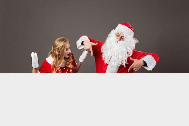 Cool Santa Claus and young beautiful mrs. Claus in sunglasses stand behind a white canvas