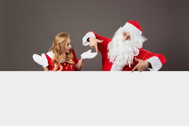 Cool Santa Claus and young beautiful mrs. Claus in sunglasses stand behind a white canvas