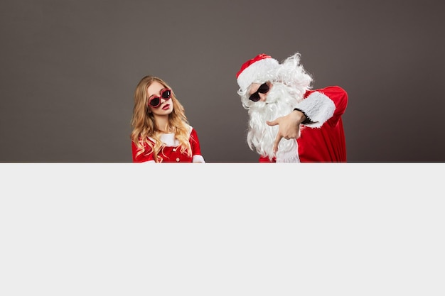 Cool Santa Claus and young beautiful mrs. Claus in sunglasses stand behind a white canvas