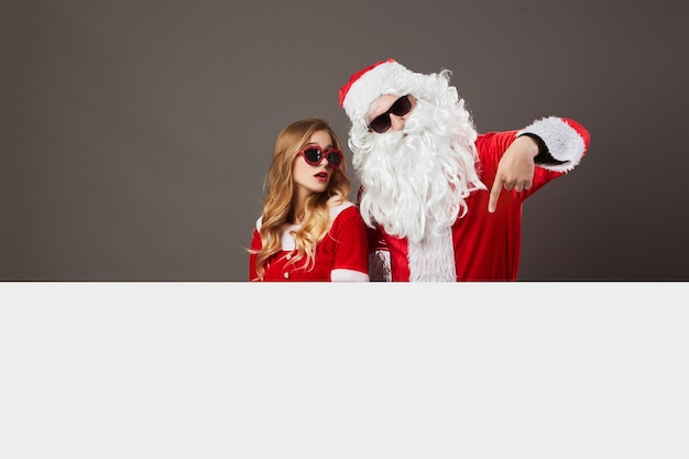 Cool Santa Claus and young beautiful mrs. Claus in sunglasses stand behind a white canvas