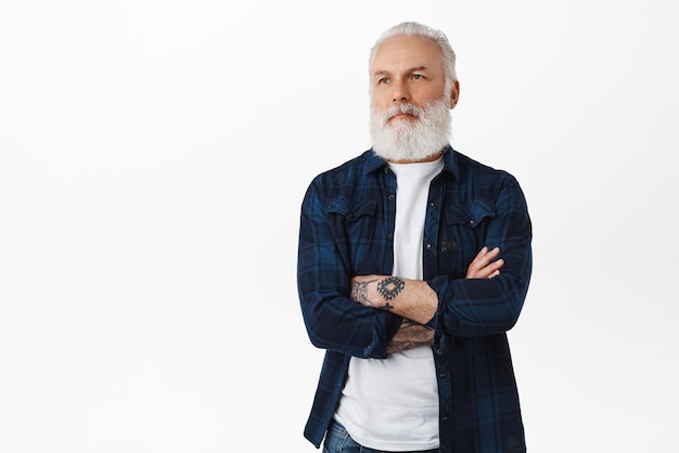 Cool senior man with tattoos bearded granfather cross arms on chest looking aside with serious cool expression standing like professional against white background