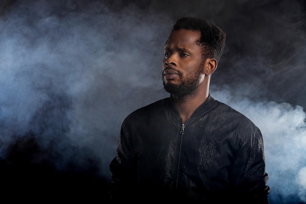 Cool strong handsome young african man with beard wearing black windbreaker jacket looking away with serious expression posing against white smoke on dark background. Copy space.