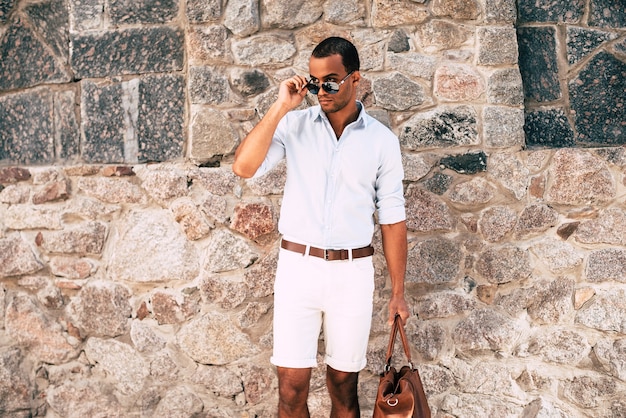 Cool and stylish. Confident young African man in smart casual clothes carrying bag and looking away while adjusting his sunglasses and standing against the stoned wall outdoors