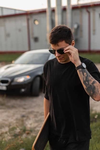 Cool stylish young man hipster with a tattoo on arm wearing a black Tshirt with a longboard walks and puts on fashion sunglasses