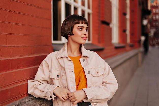 Cool woman in pink denim jacket and orange sweatshirt poses outside near brick building Attractive girl walks outdoors