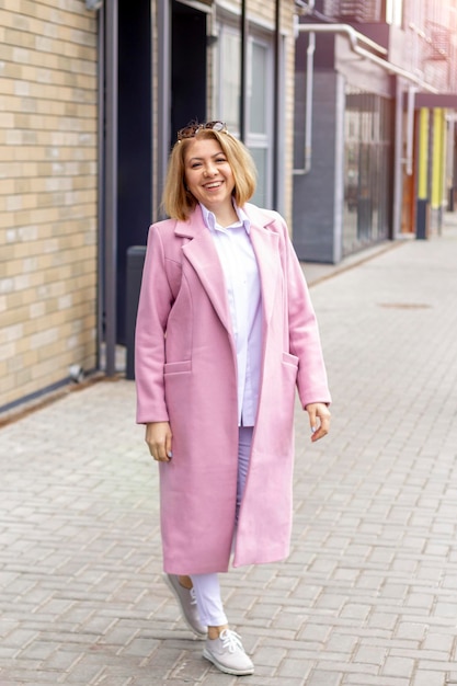 A cool young attractive woman in a pink coat and white clothes is standing on the street in the city and smilingfashion style trend black friday sale