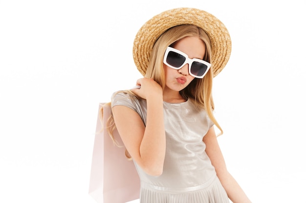 Cool young blonde girl in dress, straw hat and sunglasses holding packages on white