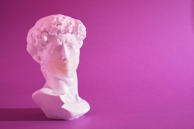 Copy of the head of an antique statue of David with a taped mouth in pink neon light on a purple background