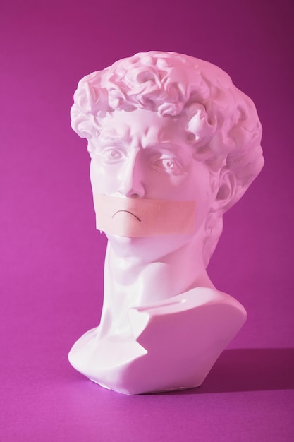 Copy of the head of an antique statue of David with a taped mouth in pink neon light on a purple background