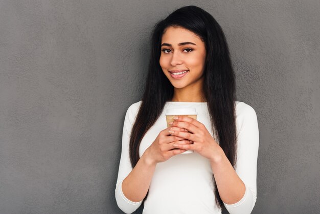 Copy space in her hand. Attractive young African woman holding coffee cup and looking at camera with smile while standing against grey background