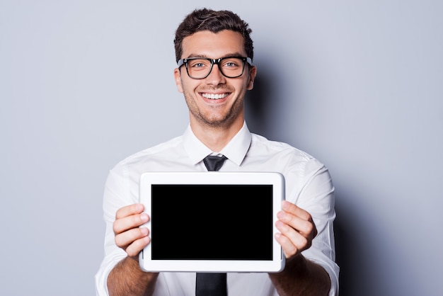 Copy space on his tablet. Handsome young man in shirt and tie showing his digital tablet and smiling while standing against grey background