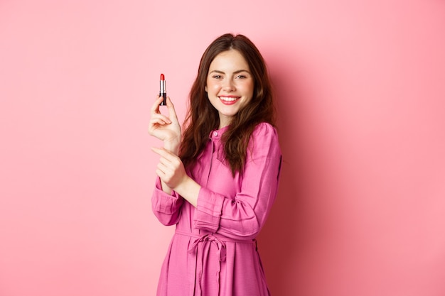 Coquettish smiling woman showing lipstick, applying make up for party event, standing in dress against pink wall.