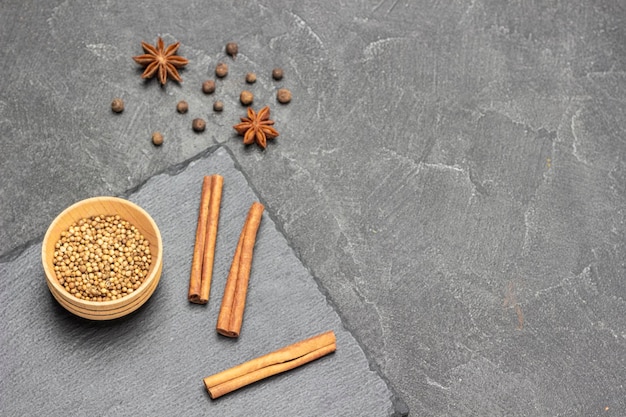 Coriander seeds in wooden bowl and cinnamon sticks on stone board Star anise and allspice on table Copy space Top view Black background