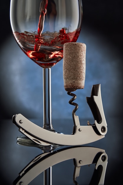 A corkscrew with a twisted wooden cork on the background of a glass of pouring red wine.