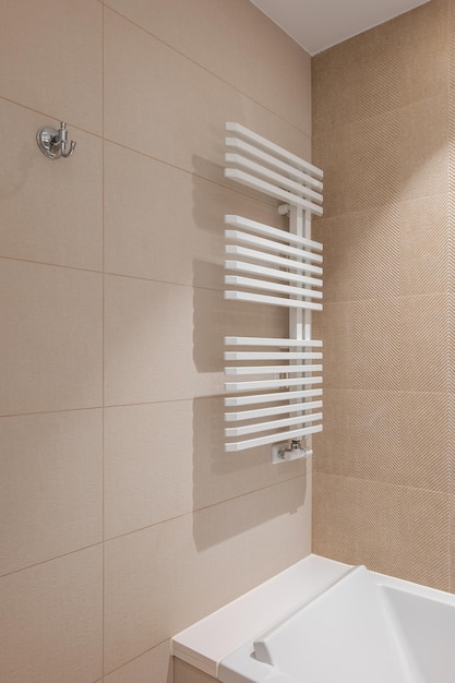 Corner of bathroom in the center with a white heating radiator on the wall for drying towels The walls of the room are lined with beige tiles of different colors Pure white acrylic bathtub